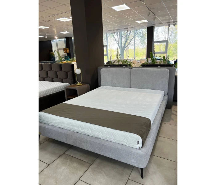Сіре ліжко на високих металевих ніжках з подвійним м’яким узголів’ям-подушками
